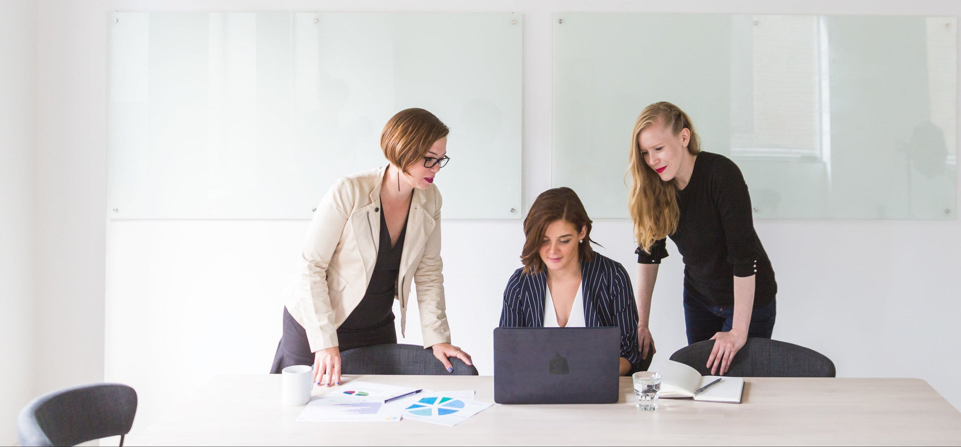 Three women working together at a table with a laptop in an office setting.